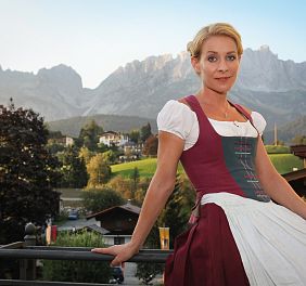 Frau in Dirndl sitzt auf Geländer vor Bergkulisse