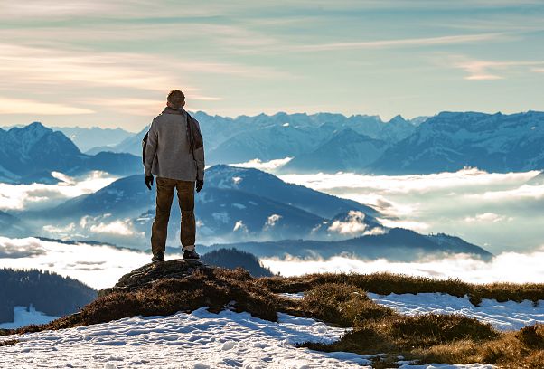 Mann steht mit Rücken zur Kamera auf Stein und blickt ins Tal