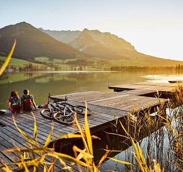 Zwei Menschen sitzen mit Rädern nebenbei auf Steeg und blicken in den Sonnenuntergang