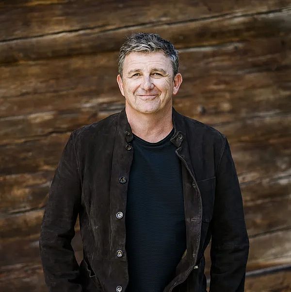 Ein Mann steht lächelnd vor einer Holzwand. Er trägt eine dunkle Jacke über einem schwarzen T-Shirt. Der Hintergrund ist verschwommen.