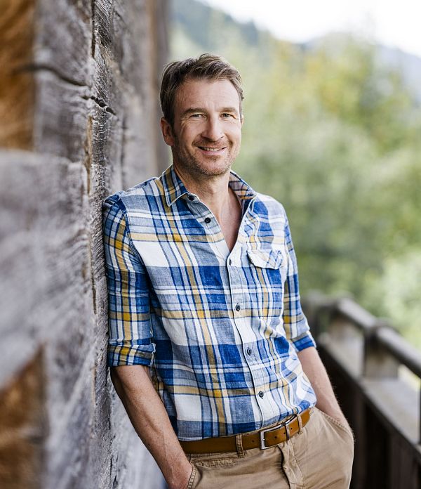 Mann in kariertem Hemd lehnt sich entspannt an eine Holzwand im Freien, lächelt und genießt die sonnige, natürliche Umgebung.