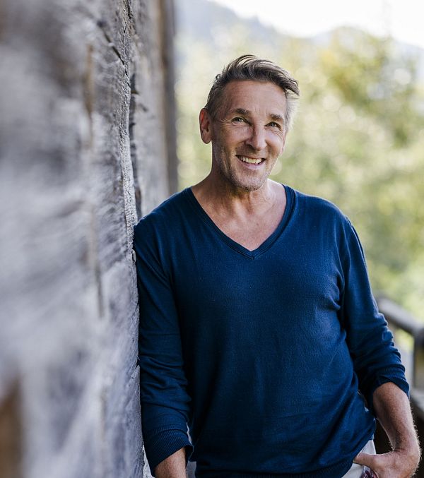 Eine lächelnde Person lehnt an einer Holzwand, umgeben von Natur im Hintergrund. Sie trägt einen blauen Pullover und wirkt entspannt und zufrieden.