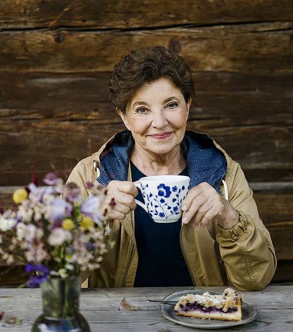 Ältere Frau hält eine Tasse mit blauem Blumenmuster und lächelt in die Kamera. Der Hintergrund ist eine Holzwand, im Vordergrund sind Blumen zu sehen.