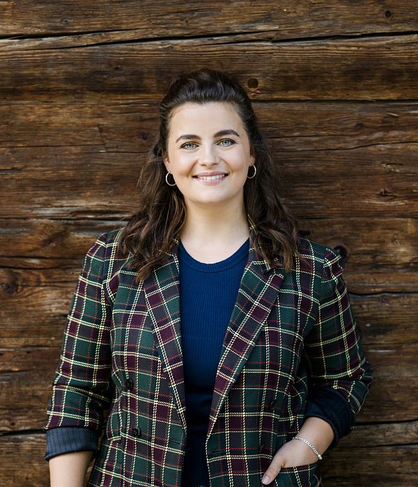 Porträt einer lächelnden Frau vor einer rustikalen Holzwand. Sie trägt einen karierten Blazer und ein dunkelblaues Oberteil. Ihre braunen Haare sind gelockt.