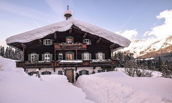 A traditional wooden chalet covered in snow, surrounded by a winter landscape with pine trees and mountains under a clear blue sky.