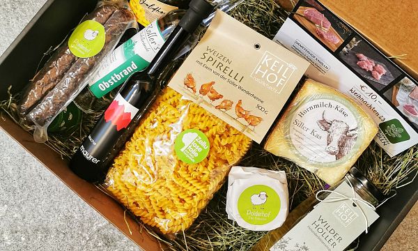 A wooden box filled with Tyrolean gourmet products from the Wilder Kaiser region, including pasta, cheese, sausages, and beverages, set on a hay-covered base.