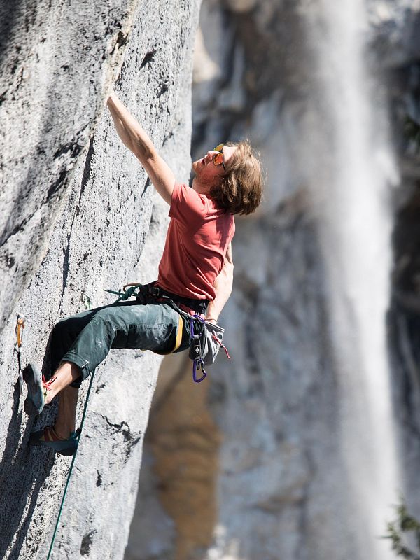 Ein Kletterer in rotem Shirt erklimmt eine steile Felswand bei Tageslicht. Sicherheitsseil und Kletterschuhe sind sichtbar. Der Hintergrund ist unscharf.