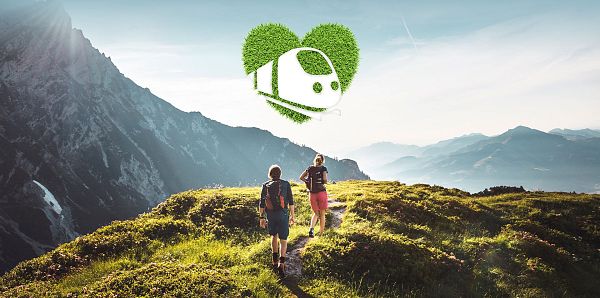 Zwei Wanderer auf einem Bergpfad, umgeben von üppiger grüner Natur und einem Bergpanorama. Ein Zug in Form eines Herzlogos symbolisiert umweltfreundliches Reisen.