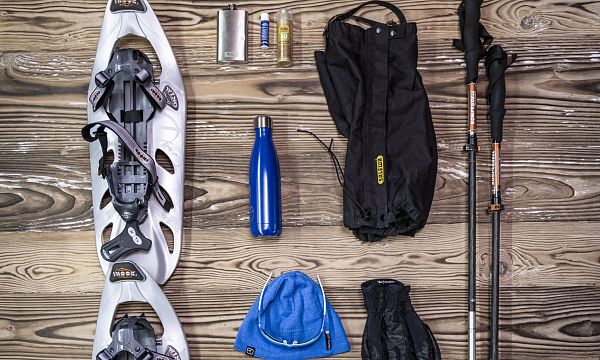 Winter hiking gear including snowshoes, poles, gloves, a blue hat, hiking boots, a water bottle, and a small snack pack on a wooden surface.