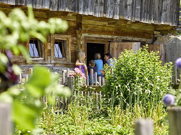 Drei Personen stehen vor einer rustikalen Holzhütte mit Garten im Vordergrund. Die Szene wirkt idyllisch und traditionell, typisch für eine ländliche Umgebung.
