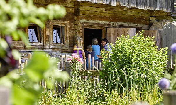 Drei Personen stehen vor einer rustikalen Holzhütte mit Garten im Vordergrund. Die Szene wirkt idyllisch und traditionell, typisch für eine ländliche Umgebung.