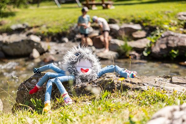 A colorful plush spider toy on a rock by a stream, with children playing nearby in the Wilder Kaiser region. Sunny day in a lush, green outdoor setting.