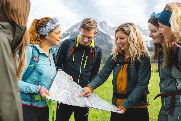Eine Gruppe von Wanderern studiert eine Karte in einer Berglandschaft. Hinter ihnen erheben sich schneebedeckte Gipfel und der Himmel ist teilweise bewölkt.