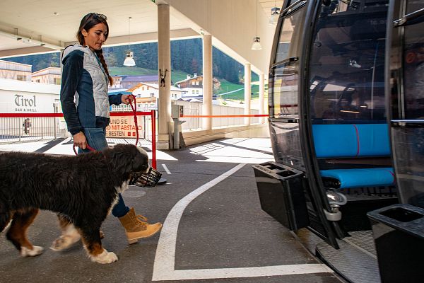 A woman with a large dog is about to enter a cable car at a station in the Wilder Kaiser region, with Tyrolean-style buildings and mountains visible.