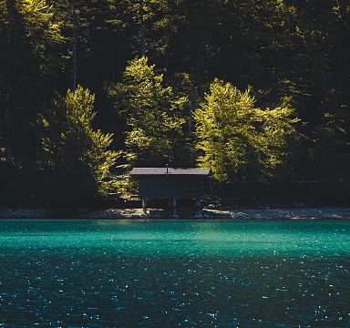 A tranquil lake with a turquoise hue, bordered by dense forest. A small wooden hut is nestled near the shoreline, under dappled sunlight filtering through the trees.