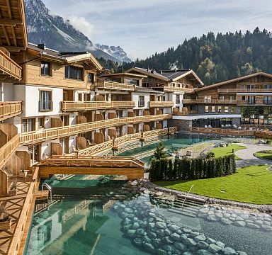 View of a luxury resort in the Wilder Kaiser, featuring traditional wooden architecture, a crystal-clear alpine pool, and mountainous backdrop on a sunny day.