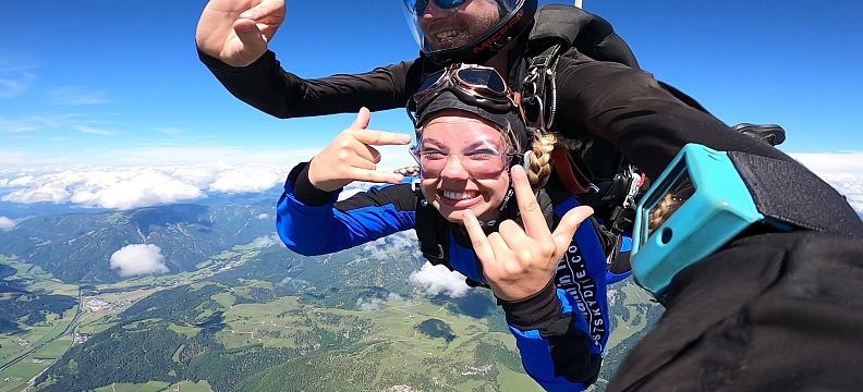 Zwei Personen beim Tandem-Fallschirmsprung hoch über einer grünen Landschaft. Sie lächeln und zeigen Handzeichen, während sie durch den blauen Himmel fallen.