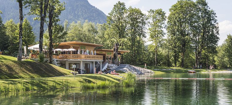 A tranquil scene at Wilder Kaiser with a lake reflecting trees and mountains, a wooden structure nearby, set against a backdrop of lush greenery.