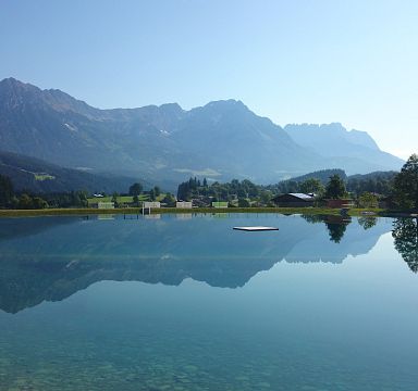 Scenic photo of the Wilder Kaiser mountains mirrored in a calm lake, surrounded by greenery and under a clear blue sky, offering a peaceful and picturesque scene.