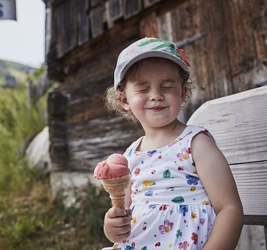 Ein kleines Mädchen in einem geblümten Kleid sitzt auf einer Bank, genießt ein Eis und lächelt verträumt mit geschlossenen Augen vor einer Holzhütte.