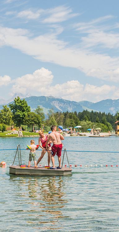 Zwei Personen stehen auf einem Floß in einem See mit Bergkulisse im Hintergrund. Der Himmel ist teils wolkig, umgeben von grünen Bäumen und Wiesen.