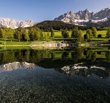 Klarer Bergsee spiegelt schneebedeckte Gipfel des Wilden Kaisers im Frühling. Saftige Wiesen und grüne Wälder umgeben die ruhige Landschaft.