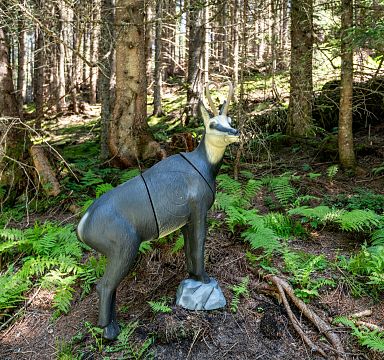 Eine plastische Gämse steht zwischen Bäumen im Wald auf grünem Boden mit Farnen. Das Holz schaut natürlich und ruhig aus, ideal für einen Spaziergang.