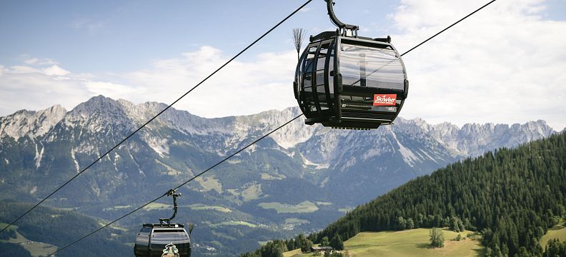 Two cable cars traveling over green hills with the imposing Wilder Kaiser mountains in the background, under a clear blue sky.