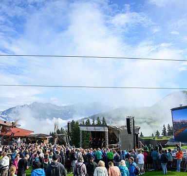 A lively outdoor scene with a crowd and stage set against the Wilder Kaiser mountains, partially veiled in mist under a clear blue sky.