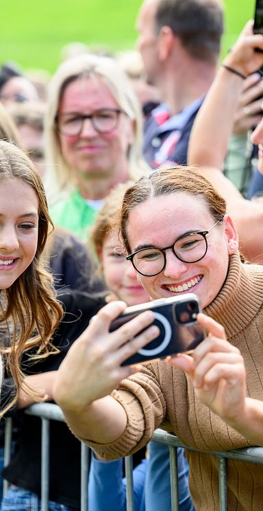 A large group of smiling people outdoors, taking photos and enjoying a gathering, with greenery in the background, suggesting a lively event atmosphere.