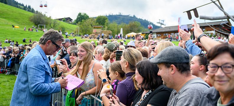 A large crowd engages with a speaker at an outdoor event in the Wilder Kaiser region, with green hills and cable cars visible in the background.