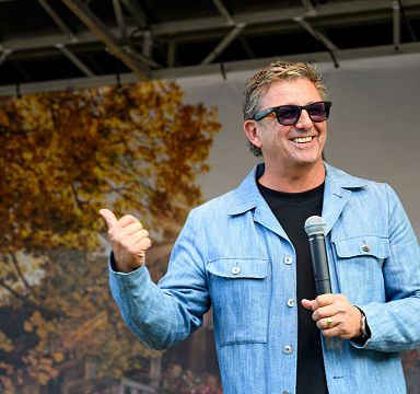 A man in sunglasses and a blue jacket holds a microphone and smiles, gesturing with his thumb, in front of a backdrop with autumn leaves.