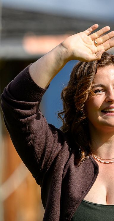 A smiling woman waves at the camera with a person filming her in the background, set outdoors on a sunny day.