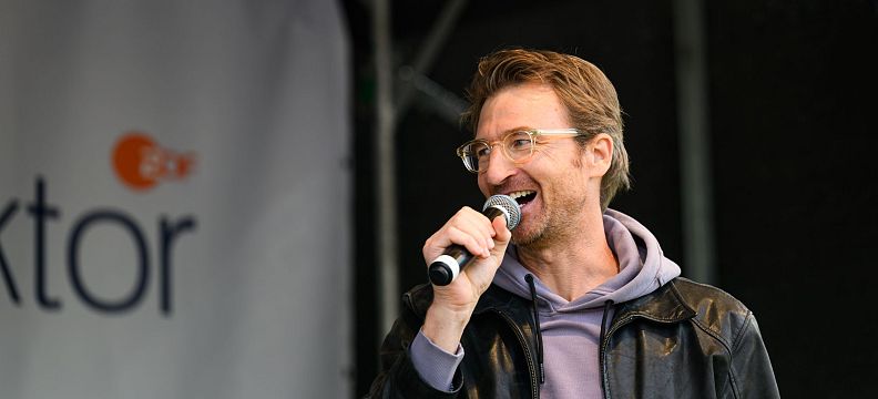 A person wearing glasses and a leather jacket speaks into a microphone on stage, smiling next to a banner with partial text visible.