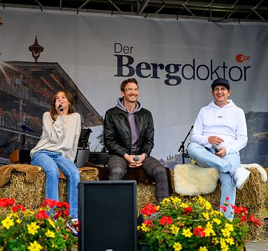 Three individuals sitting on stage at an outdoor event, surrounded by colorful flowers and rustic alpine decor, in front of a banner that reads 'Der Bergdoktor.'