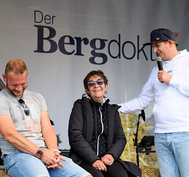 Three people are sitting on stage during an event. A man in a white hoodie is speaking into a microphone, while the other two listen and smile.