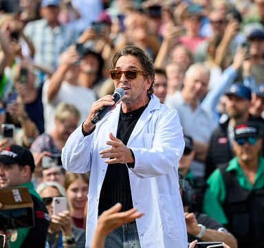 A person wearing sunglasses and a white coat speaks into a microphone in front of a crowd at an outdoor event.