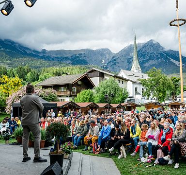 Outdoor-Veranstaltung in den Alpen mit einem Redner auf der Bühne und Publikum vor malerischer Bergkulisse.