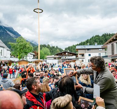 Eine Menschenmenge auf einem Dorfplatz bei einem Event. Im Hintergrund sind ein spitzer Kirchturm und bewaldete Berge zu sehen.
