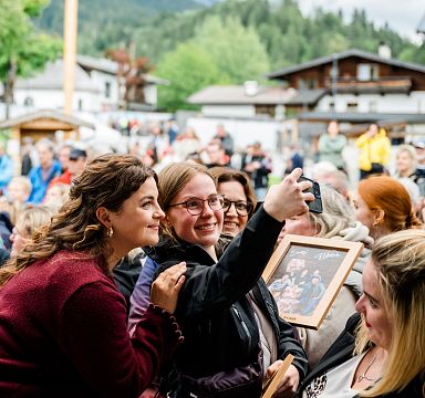Menschenmenge bei einem Freiluft-Event, während eine Gruppe von Frauen ein Selfie macht. Im Hintergrund sind alpenländische Gebäude zu sehen.