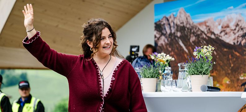 Eine Person in einem roten Oberteil winkt auf einer Bühne mit Alpenpanorama im Hintergrund. Auf dem Tisch stehen Blumen und Wassergläser.