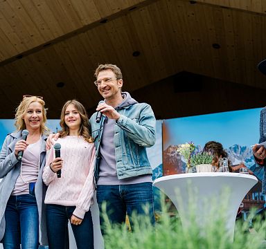 Eine Gruppe von Menschen, die auf einer Bühne stehen und Mikrofone halten, während sie lächeln. Im Hintergrund sind Bergbilder als Kulisse zu sehen.