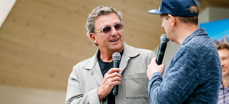 Zwei Männer auf einer Bühne unterhalten sich mit Mikrofonen. Einer trägt eine Sonnenbrille und eine helle Jacke, der andere eine blaue Kappe und Jacke.