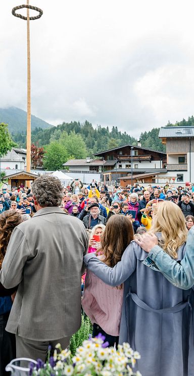 Eine große Menschenmenge auf einem Dorfplatz, umgeben von Bergen. Im Vordergrund umarmen sich mehrere Personen und schauen in die Menge.