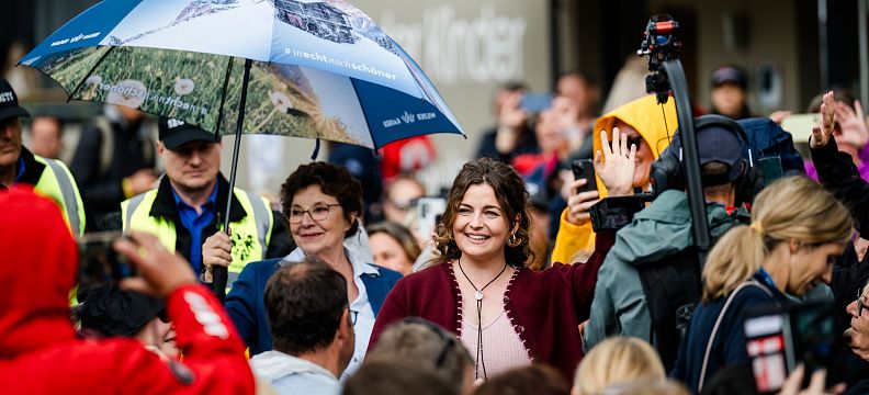 Eine Menschenmenge bei einem Event im Freien. Eine Person hält einen Regenschirm, während andere freundlich in die Kamera lächeln. Es herrscht eine fröhliche Stimmung.