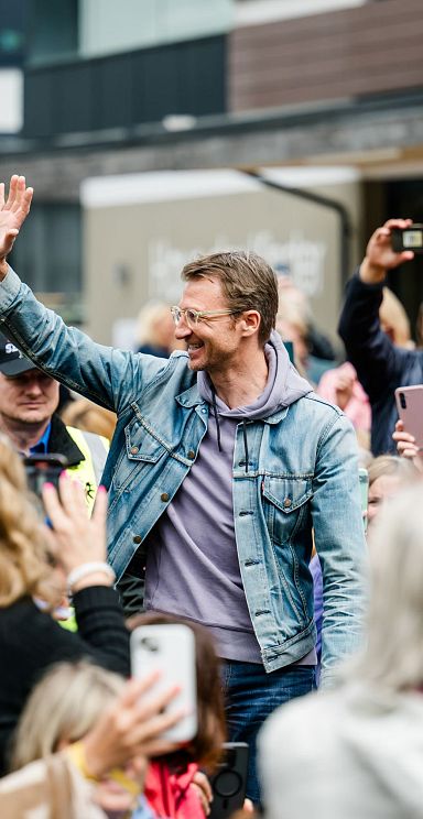 Eine Person in einer Menschenmenge, die freudig winkt. Viele umstehende Menschen halten Smartphones hoch, um Fotos oder Videos aufzunehmen.