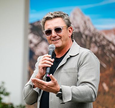 Mann mit Sonnenbrille und Mikrofon spricht auf einer Bühne, im Hintergrund unscharfe Berglandschaft.