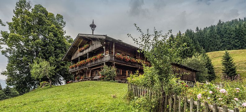 Traditionelles Tiroler Bauernhaus mit Holzfassade und Balkonblumen vor einem grünen Hang, umgeben von Bäumen und einem Holzzaun, unter bewölktem Himmel.