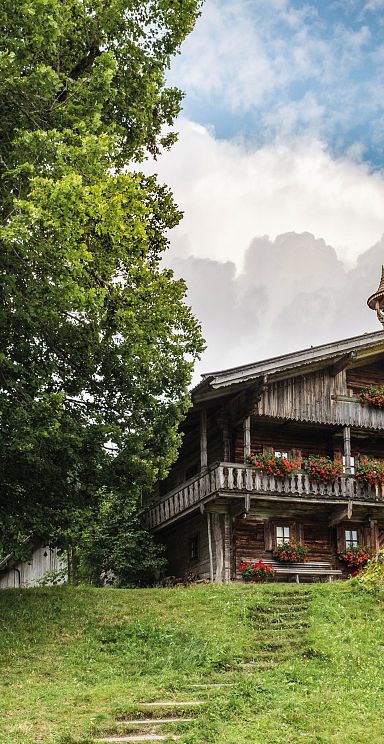 Holzhütte in Berglandschaft mit blauem Himmel, großen Bäumen und grüner Wiese. Traditionelle Architektur mit Blumen geschmücktem Balkon.