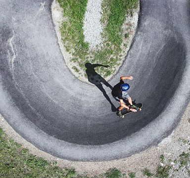 Luftaufnahme eines Radfahrers in einer engen Kurve auf einem asphaltierten Pumptrack, umgeben von grüner Vegetation und Kies.